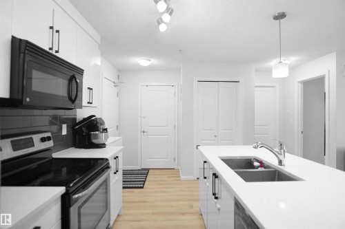 The kitchen features white cabinetry with black hardware, a black over-the-range microwave, and a stainless steel stove - 103 3353 16A Avenue, Edmonton, AB - Indoor Photo Showing Kitchen With Double Sink