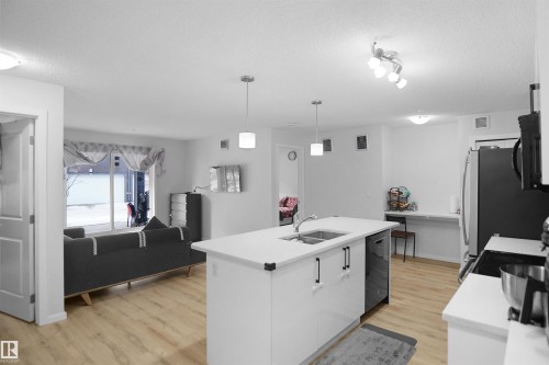 Open concept living space featuring a modern kitchen island with a double sink, light-toned flooring, and a sliding glass door leading to the exterior - 103 3353 16A Avenue, Edmonton, AB - Indoor Photo Showing Kitchen With Double Sink