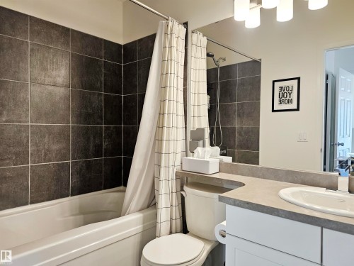 Bathroom featuring a bathtub with dark tiled surround, a toilet, a vanity with a white countertop, and a mirror with overhead lighting - 6888 Evans Wynd, Edmonton, AB - Indoor Photo Showing Bathroom