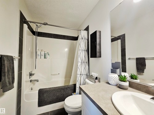 The bathroom features a bathtub and shower combination with dark tiled accents, a vanity with a countertop sink, and a large mirror - 6888 Evans Wynd, Edmonton, AB - Indoor Photo Showing Bathroom