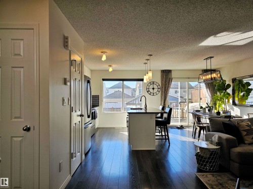 This open-concept living area features dark wood-style flooring, a kitchen island with pendant lighting, and a dining area with a view of the outdoors through a sliding glass door - 6888 Evans Wynd, Edmonton, AB - Indoor