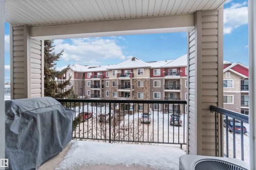 The property features a covered balcony with a black metal railing, offering views of the surrounding apartment buildings and a paved parking area - 319 17415 99 Avenue, Edmonton, AB - Outdoor With Exterior