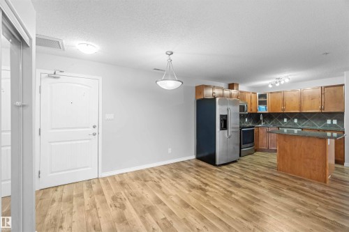 The property features an open living area with wood-style flooring, leading to a kitchen equipped with wood cabinetry, a stainless steel refrigerator, and a kitchen island with a dark countertop - 319 17415 99 Avenue, Edmonton, AB - Indoor Photo Showing Kitchen