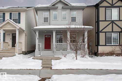 The property features a two-story exterior with light-colored siding, a covered front porch with white railings, and a distinctive red front door - 7718 Eifert Crescent, Edmonton, AB - Outdoor With Facade