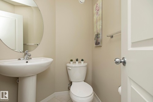 This well-appointed powder room features a pedestal sink with a chrome faucet, a coordinating toilet, and a circular wall mirror - 7718 Eifert Crescent, Edmonton, AB - Indoor Photo Showing Bathroom