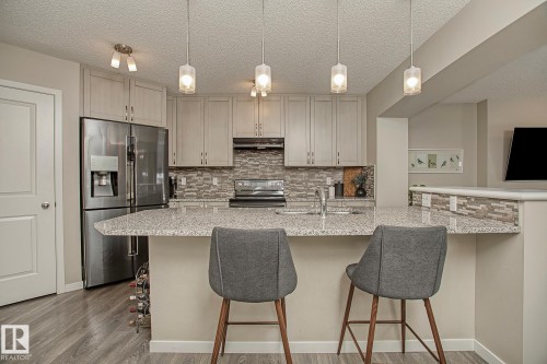 The kitchen features light-toned cabinetry, stainless steel appliances, a tiled backsplash, and an island with a granite countertop and pendant lighting - 7718 Eifert Crescent, Edmonton, AB - Indoor Photo Showing Kitchen With Upgraded Kitchen
