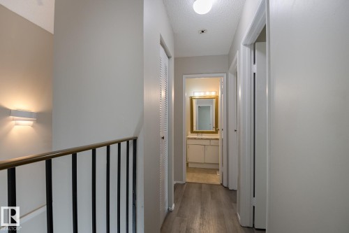 Hallway with light-colored walls, wood-style flooring, a wall sconce, and a glimpse of a bathroom with a vanity and mirror - 37 Woodvale Village, Edmonton, AB - Indoor Photo Showing Other Room