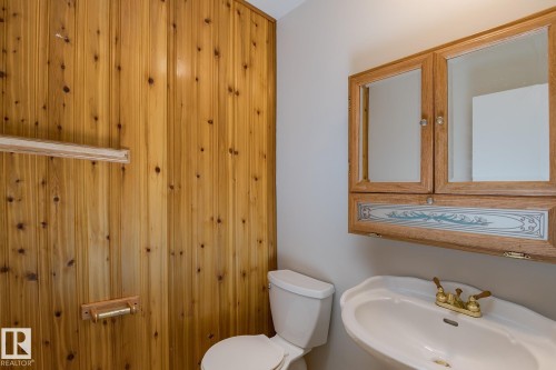 This bathroom features wood paneling, a wall-mounted sink with brass-toned fixtures, a toilet, and a wall-mounted wooden cabinet with mirrored doors - 37 Woodvale Village, Edmonton, AB - Indoor Photo Showing Bathroom
