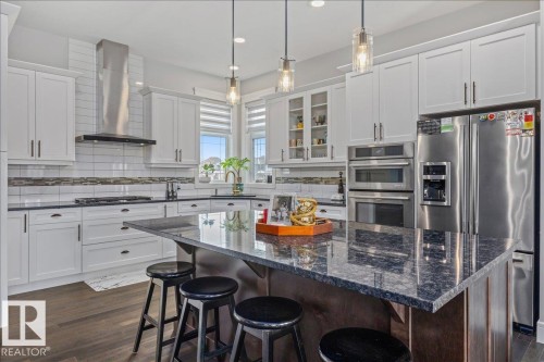The kitchen features white cabinetry, dark countertops, and stainless steel appliances including a refrigerator, double oven, and range hood - 3908 Ginsburg Crescent, Edmonton, AB - Indoor Photo Showing Kitchen With Upgraded Kitchen