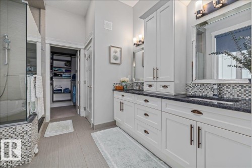 Bathroom featuring a double vanity with white cabinetry, dark countertops, and a tiled backsplash, alongside a shower with a glass enclosure and mosaic tile detailing - 3908 Ginsburg Crescent, Edmonton, AB - Indoor Photo Showing Other Room