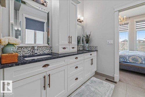This bathroom features a double vanity with white cabinetry, dark countertops, and a mosaic tile backsplash - 3908 Ginsburg Crescent, Edmonton, AB - Indoor Photo Showing Bedroom