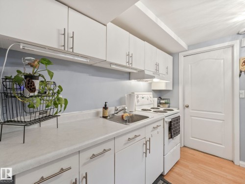 The kitchen features white cabinetry with metal handles, a white countertop, a stainless steel sink, and light wood flooring - 12141 96 Street, Edmonton, AB - Indoor Photo Showing Kitchen
