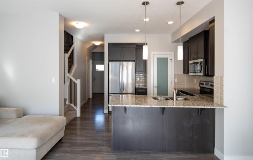 The kitchen features dark wood cabinetry, stainless steel appliances, a tiled backsplash, and a granite countertop island with a sink and pendant lighting - 1793 Keene Crescent, Edmonton, AB - Indoor Photo Showing Kitchen With Double Sink With Upgraded Kitchen