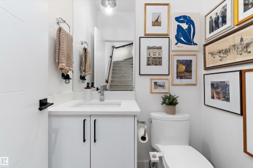 This bathroom features a contemporary white vanity with black hardware, a pristine white toilet, and a collection of framed artwork adorning the wall - 1305 Enright Landing, Edmonton, AB - Indoor Photo Showing Bathroom