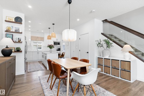 Open concept living area with light-toned flooring, a modern kitchen with white cabinetry and an island, and a staircase featuring a glass railing - 1305 Enright Landing, Edmonton, AB - Indoor
