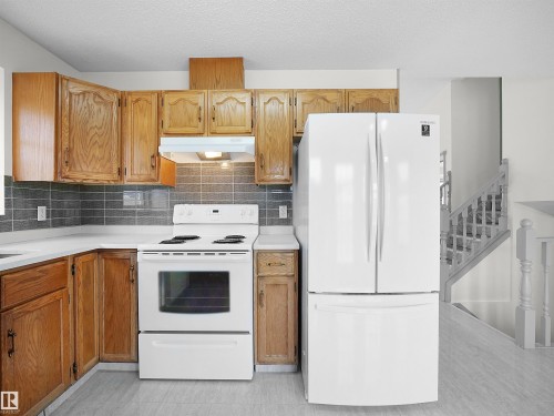 The kitchen features light wood cabinetry, white appliances, and a tiled backsplash, with a staircase visible in the background - 14523 32 Street, Edmonton, AB - Indoor Photo Showing Kitchen