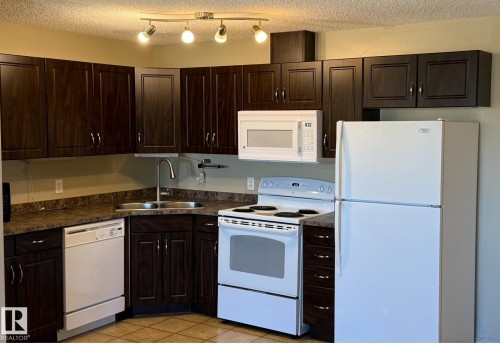 Corner kitchen featuring dark wood-finish cabinetry, white appliances, and a double basin stainless steel sink - 408 309 Clareview Station Drive Nw, Edmonton, AB 