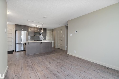 The property features an open-concept layout with light-toned flooring, leading to a kitchen area with gray cabinetry, stainless steel appliances, and a central island with counter seating - 309 4008 Savaryn Drive, Edmonton, AB - Indoor Photo Showing Kitchen With Stainless Steel Kitchen