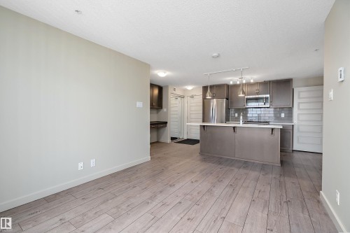 The open concept living area features light-toned flooring, with a modern kitchen showcasing stainless steel appliances, grey cabinetry, a subway tile backsplash, and an island with pendant lighting - 309 4008 Savaryn Drive, Edmonton, AB - Indoor Photo Showing Kitchen With Stainless Steel Kitchen