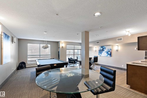 Well-lit recreational space featuring a pool table, a glass-top table with seating, and a partial view of a kitchen area with dark wood cabinetry - 309 4008 Savaryn Drive, Edmonton, AB - Indoor