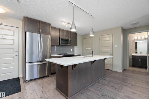 The kitchen features modern stainless steel appliances, dark wood cabinetry, and a large island with a white countertop - 309 4008 Savaryn Drive, Edmonton, AB - Indoor Photo Showing Kitchen With Stainless Steel Kitchen With Upgraded Kitchen