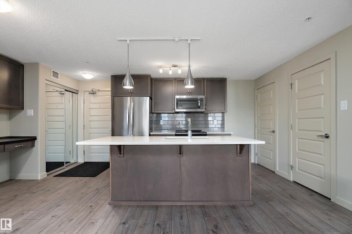 The kitchen features a central island with a white countertop and pendant lighting, stainless steel appliances, and dark cabinetry - 309 4008 Savaryn Drive, Edmonton, AB - Indoor Photo Showing Kitchen With Stainless Steel Kitchen With Upgraded Kitchen