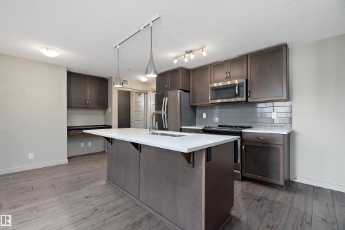 Modern kitchen featuring a large island with a white countertop, dark wood cabinetry, stainless steel appliances, and a tiled backsplash - 309 4008 Savaryn Drive, Edmonton, AB - Indoor Photo Showing Kitchen With Stainless Steel Kitchen With Upgraded Kitchen