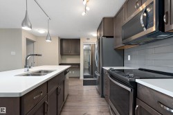 The kitchen features dark wood cabinetry, stainless steel appliances, a white countertop island with a double sink, and a gray tiled backsplash - 