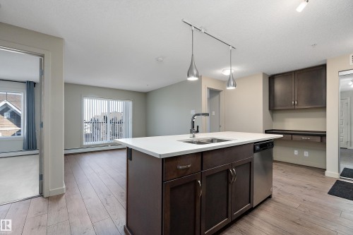 The kitchen features a central island with a sink and a stainless steel dishwasher, complemented by dark wood cabinetry and a light-colored countertop - 309 4008 Savaryn Drive, Edmonton, AB - Indoor Photo Showing Kitchen With Double Sink