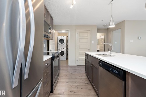 The kitchen features stainless steel appliances, dark wood cabinetry, a large central island with a sink, and light-colored countertops - 309 4008 Savaryn Drive, Edmonton, AB - Indoor Photo Showing Kitchen With Stainless Steel Kitchen With Double Sink