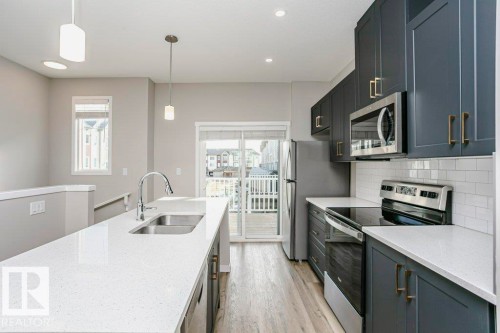 The kitchen features a spacious island with a sink, white countertops, and dark cabinetry with gold-toned hardware - 99 320 Secord Boulevard, Edmonton, AB - Indoor Photo Showing Kitchen With Stainless Steel Kitchen With Double Sink With Upgraded Kitchen
