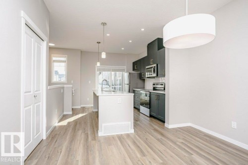 Open concept living area with light-toned flooring, featuring a modern kitchen with dark cabinetry and stainless steel appliances - 99 320 Secord Boulevard, Edmonton, AB - Indoor Photo Showing Kitchen