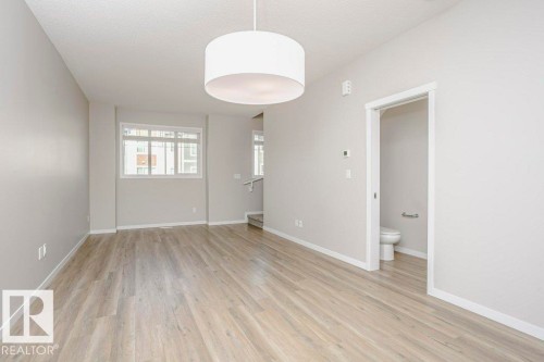 Spacious living area featuring light-toned flooring, neutral wall paint, and a modern drum pendant light fixture - 99 320 Secord Boulevard, Edmonton, AB - Indoor Photo Showing Other Room