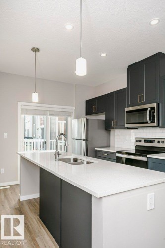 Modern kitchen featuring dark cabinetry, stainless steel appliances, a white subway tile backsplash, and an island with a light-colored countertop - 99 320 Secord Boulevard, Edmonton, AB - Indoor Photo Showing Kitchen With Stainless Steel Kitchen With Double Sink With Upgraded Kitchen