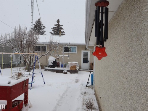 The property features a stucco exterior, a rear entrance, and a snow-covered yard with mature trees - 4315 56 Avenue, Barrhead, AB - Outdoor