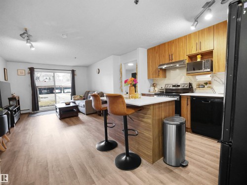 Open concept living space featuring light-toned flooring, a kitchen island with seating, and a kitchen with wood-look cabinetry and stainless steel appliances - 106 2305 35A Avenue Nw, Edmonton, AB - Indoor Photo Showing Kitchen