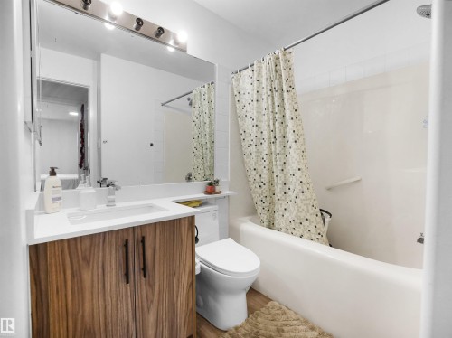 Bathroom featuring a modern vanity with wood-grain cabinetry and a white countertop, a large mirror, and a full-size bathtub with an overhead shower - 106 2305 35A Avenue Nw, Edmonton, AB - Indoor Photo Showing Bathroom