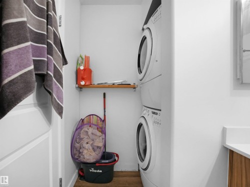 Dedicated laundry area featuring a stacked washer and dryer, a wall-mounted wooden shelf, and a white countertop - 106 2305 35A Avenue Nw, Edmonton, AB - Indoor Photo Showing Laundry Room