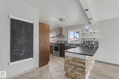 The kitchen features dark wood cabinetry, stainless steel appliances, a unique tiled backsplash, and a breakfast bar with a stone-clad base - 5920 132 Avenue, Edmonton, AB - Indoor