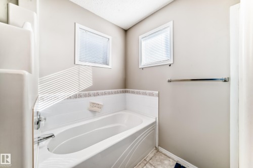 Bathroom with a built-in bathtub, two windows with blinds, and light-colored wall tiles with a decorative border - 19024 50 Avenue, Edmonton, AB - Indoor Photo Showing Bathroom