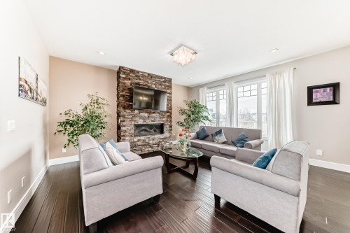 The living area features dark hardwood flooring, a prominent stone-clad fireplace with an inset television, and large windows with white trim - 4134 Charles Link, Edmonton, AB - Indoor Photo Showing Living Room With Fireplace