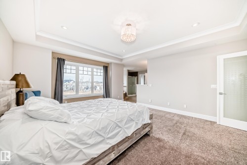 This spacious room features a tray ceiling with recessed lighting and a decorative chandelier, a large window with a view, and neutral-toned carpeting - 4134 Charles Link, Edmonton, AB - Indoor Photo Showing Bedroom
