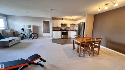 Open concept living area with light-colored carpet, a dining space, and a kitchen featuring white cabinetry, stainless steel appliances, and dark flooring - 1318 330 Clareview Station Drive Nw, Edmonton, AB - Indoor