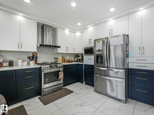 The kitchen features white upper cabinetry with brass hardware, dark blue lower cabinetry, a stainless steel range with a range hood, and a stainless steel refrigerator - 1147 Lincoln Crescent, Edmonton, AB - Indoor Photo Showing Kitchen With Upgraded Kitchen