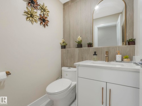 The bathroom features a modern vanity with a white countertop and gold-toned faucet, a large mirror with an arched gold frame, and vertically textured wall tiling - 1147 Lincoln Crescent, Edmonton, AB - Indoor Photo Showing Bathroom