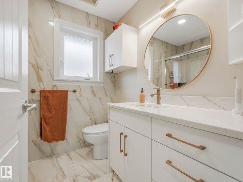 The bathroom features a modern vanity with a white countertop and gold-toned hardware, a large circular mirror with a gold-toned frame, and light-colored tiled walls and flooring - 1147 Lincoln Crescent, Edmonton, AB - Indoor Photo Showing Bathroom