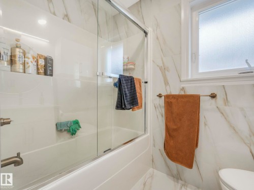 Bathroom featuring a bathtub with a sliding glass door, white wall tiling with marble-like veining, and a window with frosted glass - 1147 Lincoln Crescent, Edmonton, AB - Indoor Photo Showing Bathroom