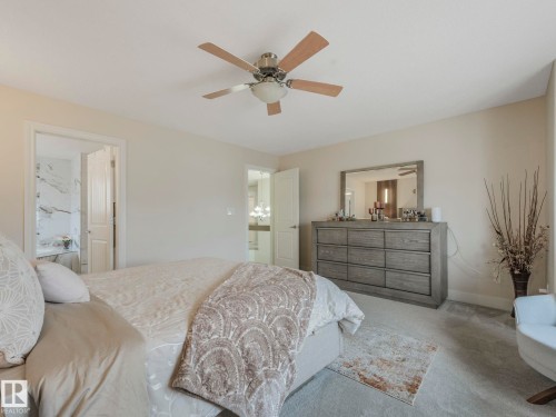 This bedroom features light-colored walls and carpeting, a ceiling fan, and offers an entryway to a bathroom with marble-patterned wall tiles - 1147 Lincoln Crescent, Edmonton, AB - Indoor Photo Showing Bedroom