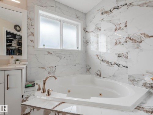 The bathroom features white marbled wall tiling, a white built-in bathtub with a gold-toned faucet, and a vanity with white cabinetry and a mirror - 1147 Lincoln Crescent, Edmonton, AB - Indoor Photo Showing Bathroom