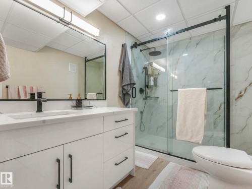 Well-appointed bathroom featuring a vanity with a white countertop and dark fixtures, a large mirror with a dark frame, and a shower with glass doors and a white marble-patterned surround - 1147 Lincoln Crescent, Edmonton, AB - Indoor Photo Showing Bathroom